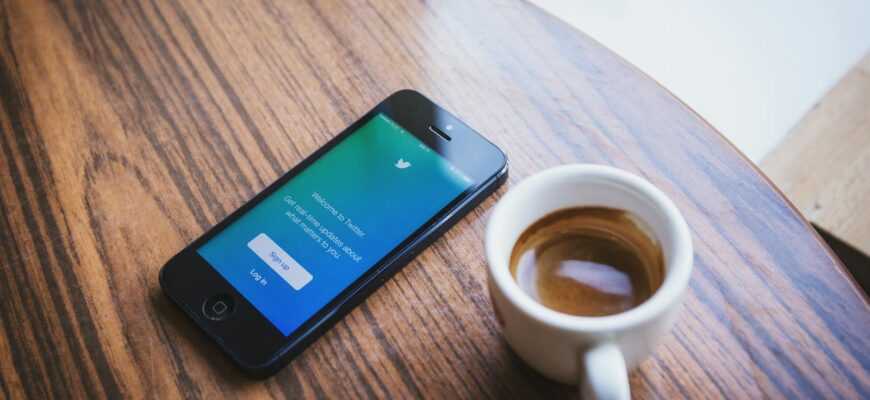 Close-up of a smartphone with the Twitter app next to a cup of coffee on a wooden table.